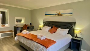 A hotel room with a large bed, a desk, a chair, a nightstand, and a painting on the wall. - Ngandu at Sea  (Walvis Bay)