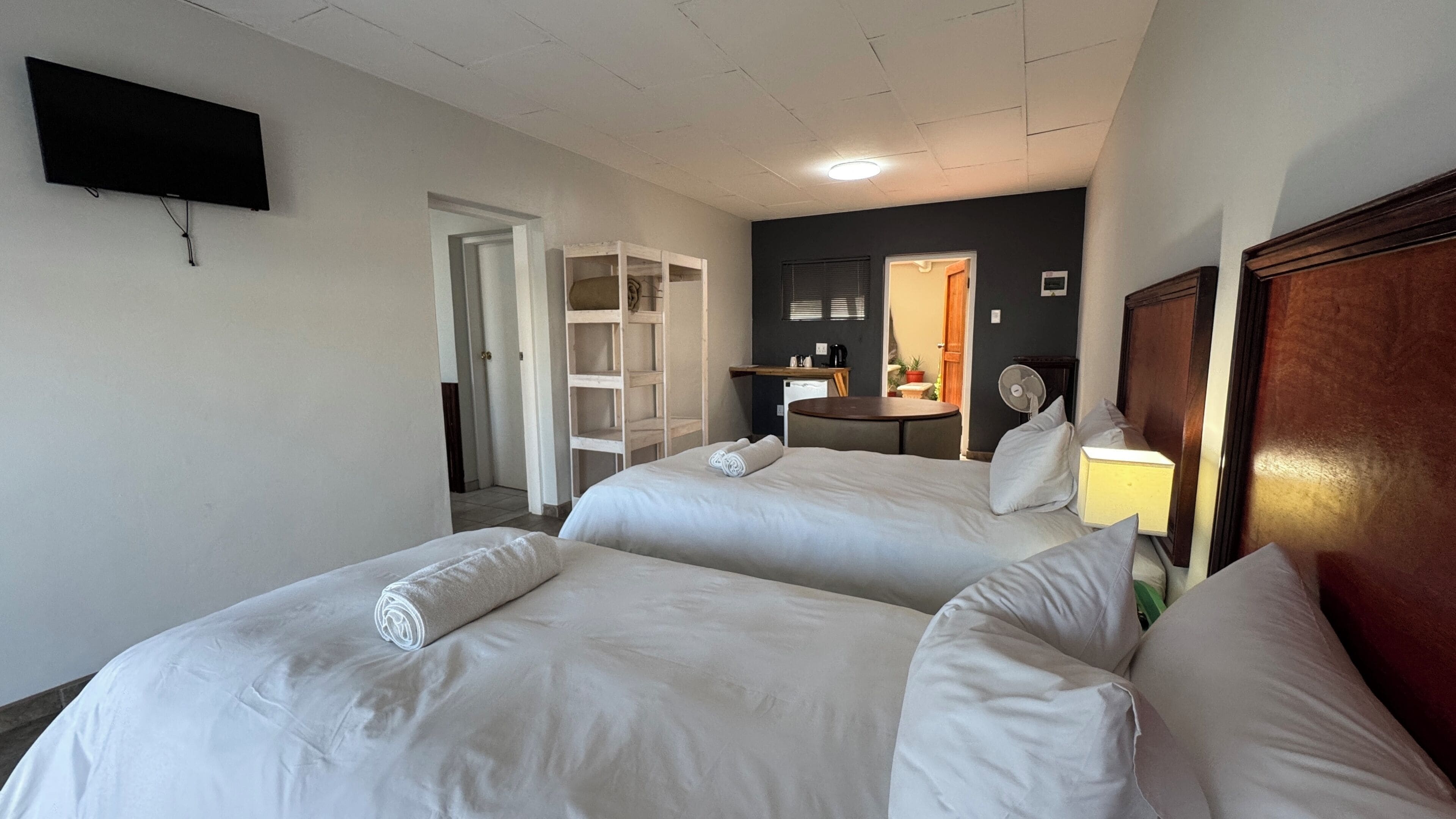 A hotel room with two beds, a television mounted on the wall, a desk with a chair, and a small table with a fan.