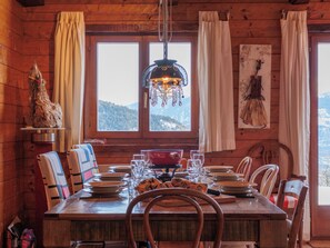 Ein Essbereich in einer Holzhütte mit einem Tisch für vier Personen, einer Pendelleuchte und Blick auf eine verschneite Berglandschaft durch ein Fenster.