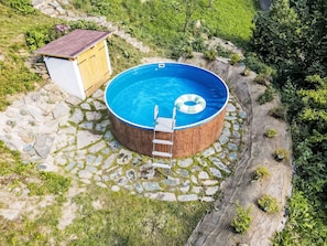Une piscine ronde, dotée d’une terrasse en bois et d’une échelle, entourée d’une bordure en pierre et d’herbe.