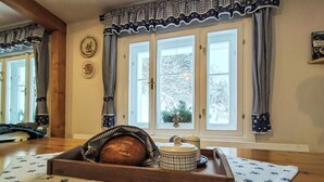 Una habitación acogedora con piso de madera, un ventanal con cortinas cuadriculadas, un plato decorativo en la pared y una cesta de pan sobre una mesa.