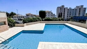 Piscina retangular com deck ladrilhado, rodeada por edifícios e vegetação.
