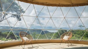 Habitación con un ventanal grande que ofrece vistas a un paisaje montañoso. Hay dos sillones de mimbre con cojines.