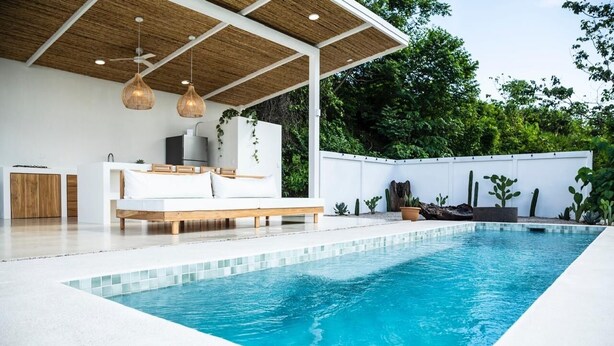 Un área de piscina moderna al aire libre con piscina blanca, terraza de madera y zona de estar cubierta con sofá e iluminación colgante.