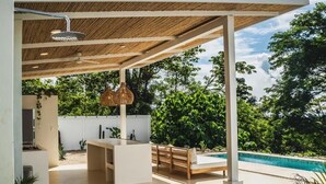 Una cocina exterior moderna con una parrilla integrada, un área de estar con un banco y una piscina al fondo.