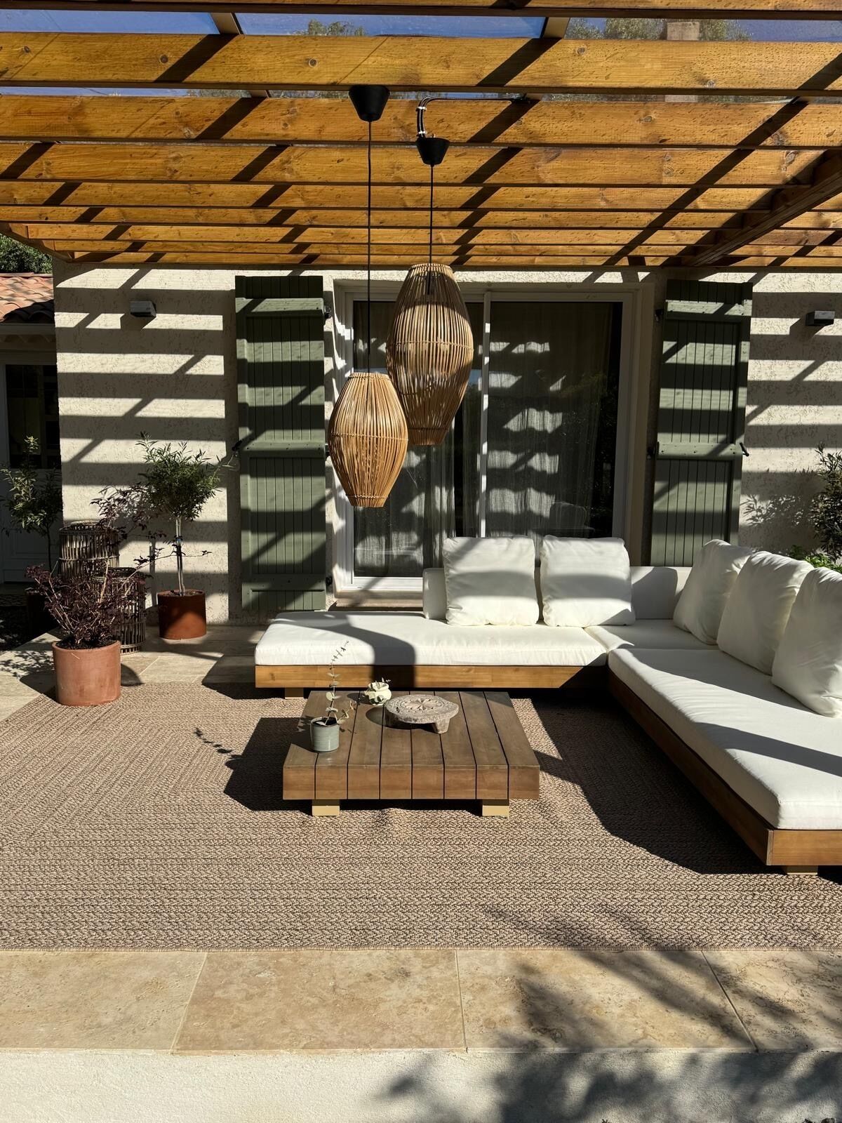 Un'area salotto esterna moderna con una pergola in legno, due lampade in vimini e un divano bianco con cuscini.
