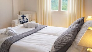Ein ordentlich bezogenes Bett mit weißen Leinen und einer grauen Decke, eine Nachttischlampe, ein kleiner Topfpflanze und ein Sonnenblumenbild an der Wand.