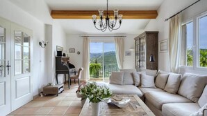 Ein geräumiges Wohnzimmer mit einer Sofagarnitur, einem Couchtisch mit Blumenvase, einem Klavier und Blick ins Freie durch große Fenster.