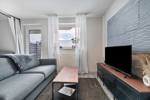 A modern living room with a grey sofa, a wooden coffee table, a flat-screen TV on a wooden stand, and a view through a window with sheer curtains. - Holiday Home 'Klein Aber Fein' with Shared Terrace and Wi-Fi (Worpswede)