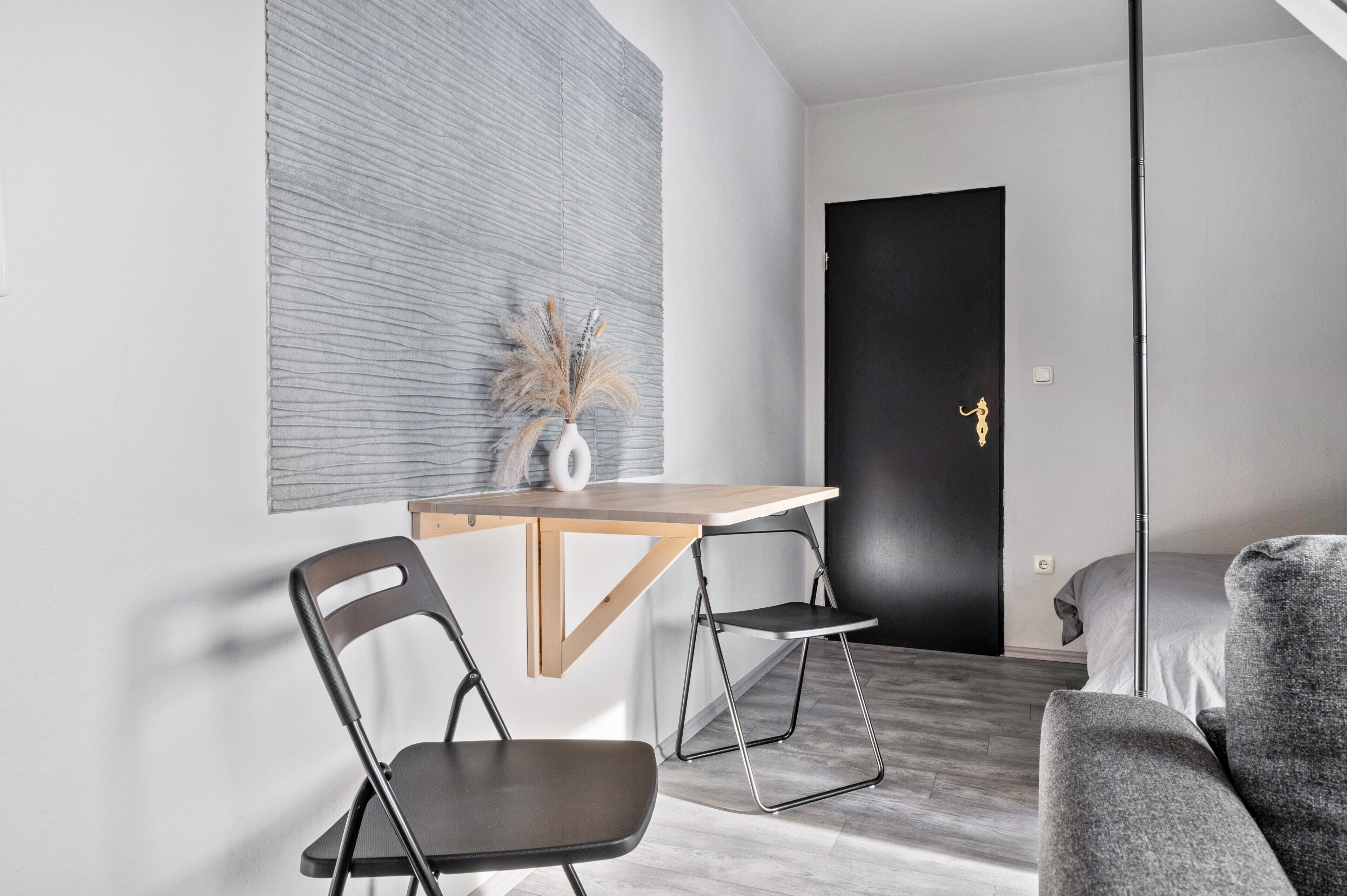 Ein modernes Zimmer mit klappbarem Schreibtisch, einem Sessel, einem Bett und einer schwarzen Tür.