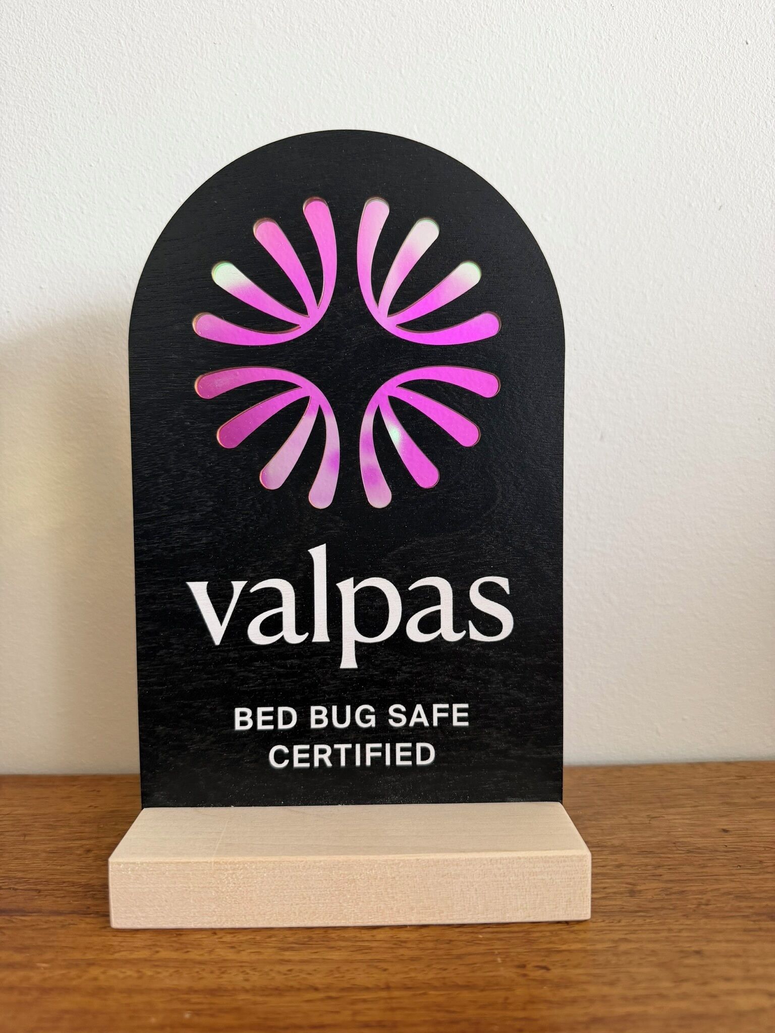 A black plaque with a pink and white logo and text stating 'valpas BED BUG SAFE CERTIFIED' on a wooden base.