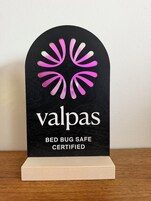 A black plaque with a pink and white logo and text stating 'valpas BED BUG SAFE CERTIFIED' on a wooden base.