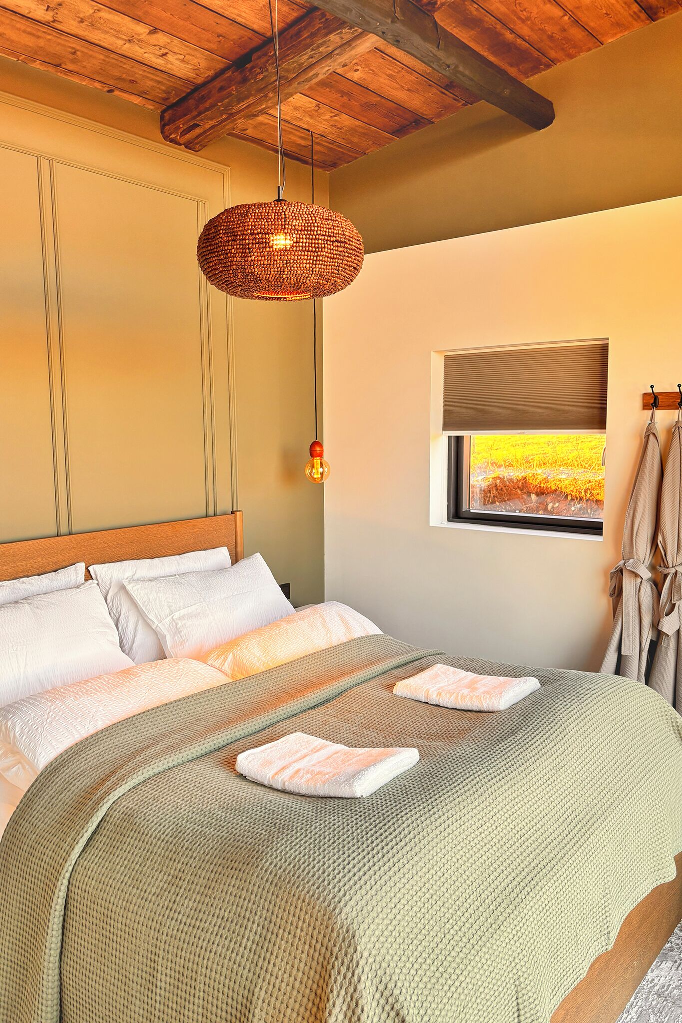 A bedroom with a wooden ceiling, a bed with white bedding, a woven light fixture, and a window with a view of a field.