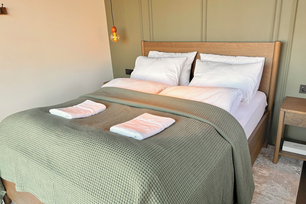 A neatly made bed with a green textured blanket and two towels, a wooden headboard, and a bedside table with a lamp.