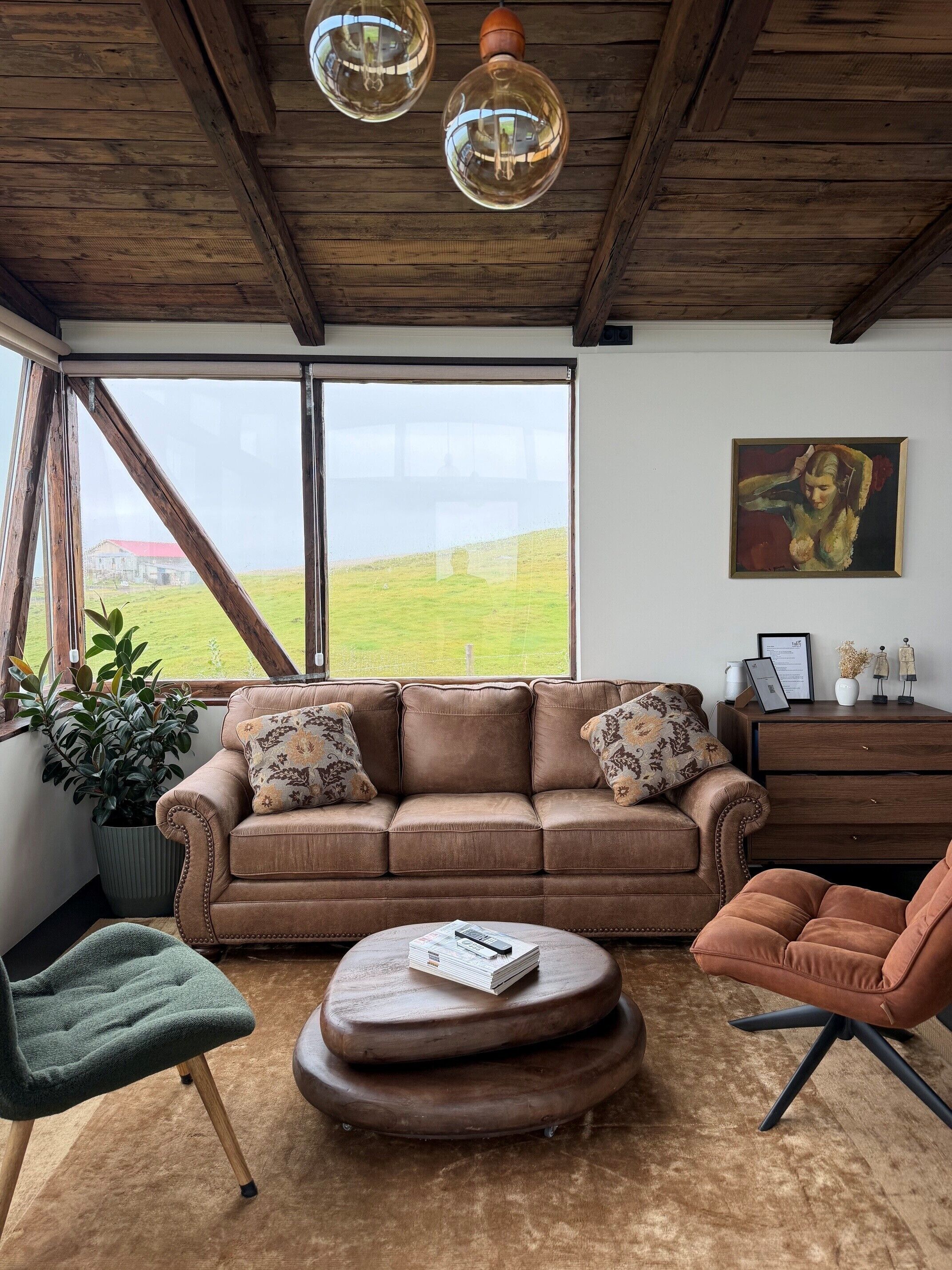 A living room with a brown leather sofa, a green chair, a wooden coffee table, and a large window with a view of a green field.