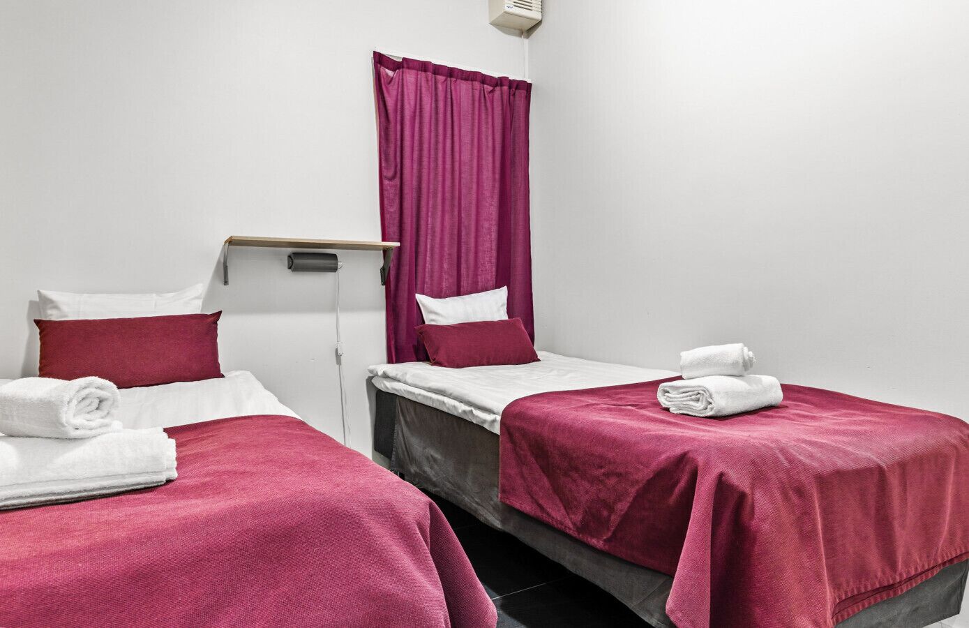 A hotel room with two single beds, each with a red bedspread and white pillows. There is a small table between the beds, and a window with purple curtains.