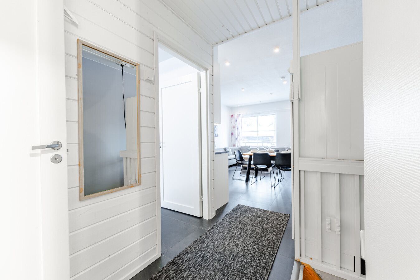 En gang med hvide vægge og et gråt tæppe, der fører til et spiseområde med et bord og stole.