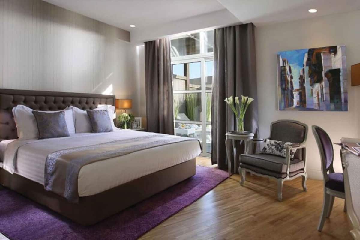 Una habitación de hotel moderna con una cama grande, una mesita pequeña, una silla y un ventanal con cortinas.