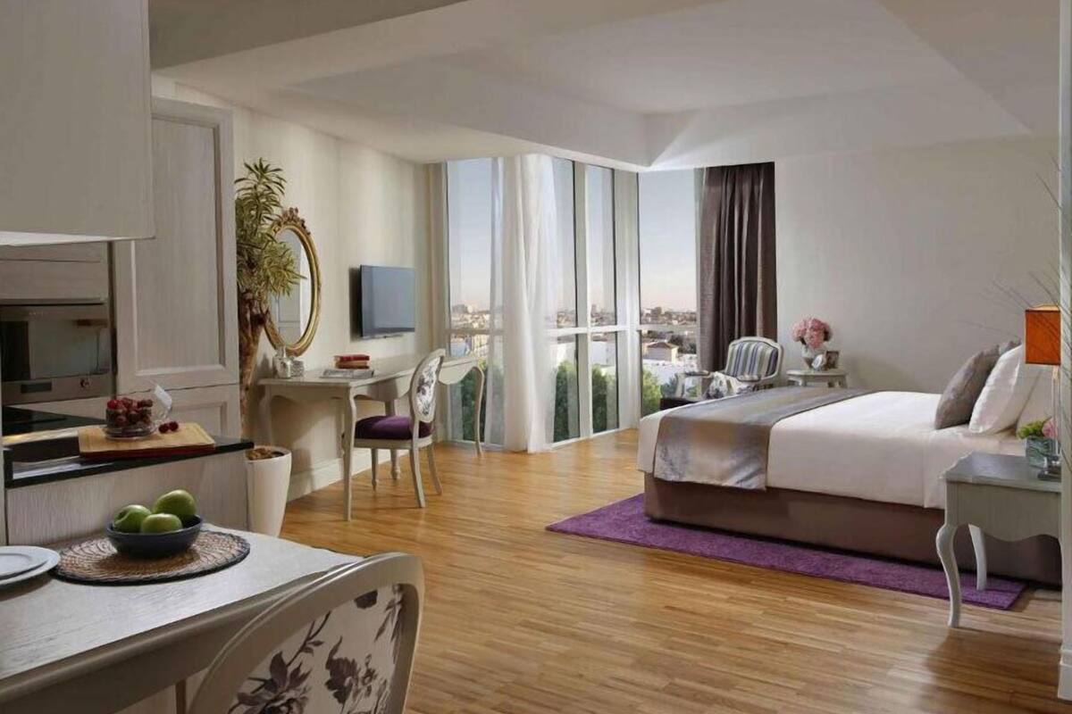 Habitación de hotel moderna con una cama grande, un escritorio con silla, televisor y vistas de la ciudad a través de amplios ventanales.