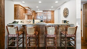 A kitchen with wooden cabinets, a marble countertop, and four wicker bar stools. - Grand Lodge - 3 Bedroom Deluxe #404 - Ski-in/out 3 Condo (Park City)