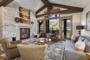 Ein gemütlicher Wohnraum mit Kamin aus Stein, Ledersofas und Blick auf eine gebirgige Landschaft durch große Fenster.