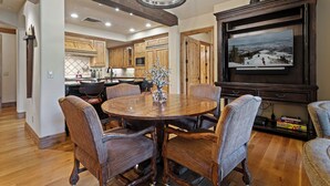 A spacious dining area with a round wooden table and six upholstered chairs, a large flat-screen TV mounted on a dark wooden cabinet, and a kitchen in the background with wooden cabinets and a tiled backsplash. - Ski-in/ski-out Deer Valley Escape 2 Bedroom Condo (Park City)