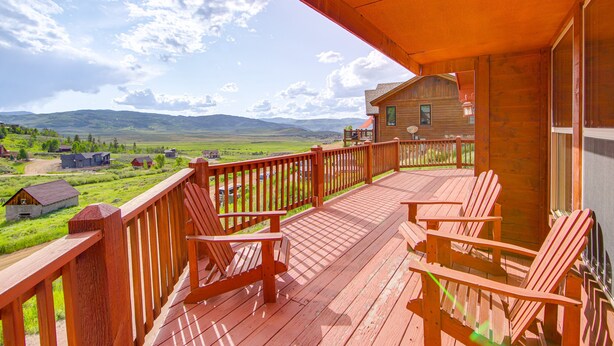 Eine Holzterrasse mit Adirondack-Stühlen, im Hintergrund ein Holzhaus und eine Aussicht auf eine grüne Landschaft mit Hügeln.