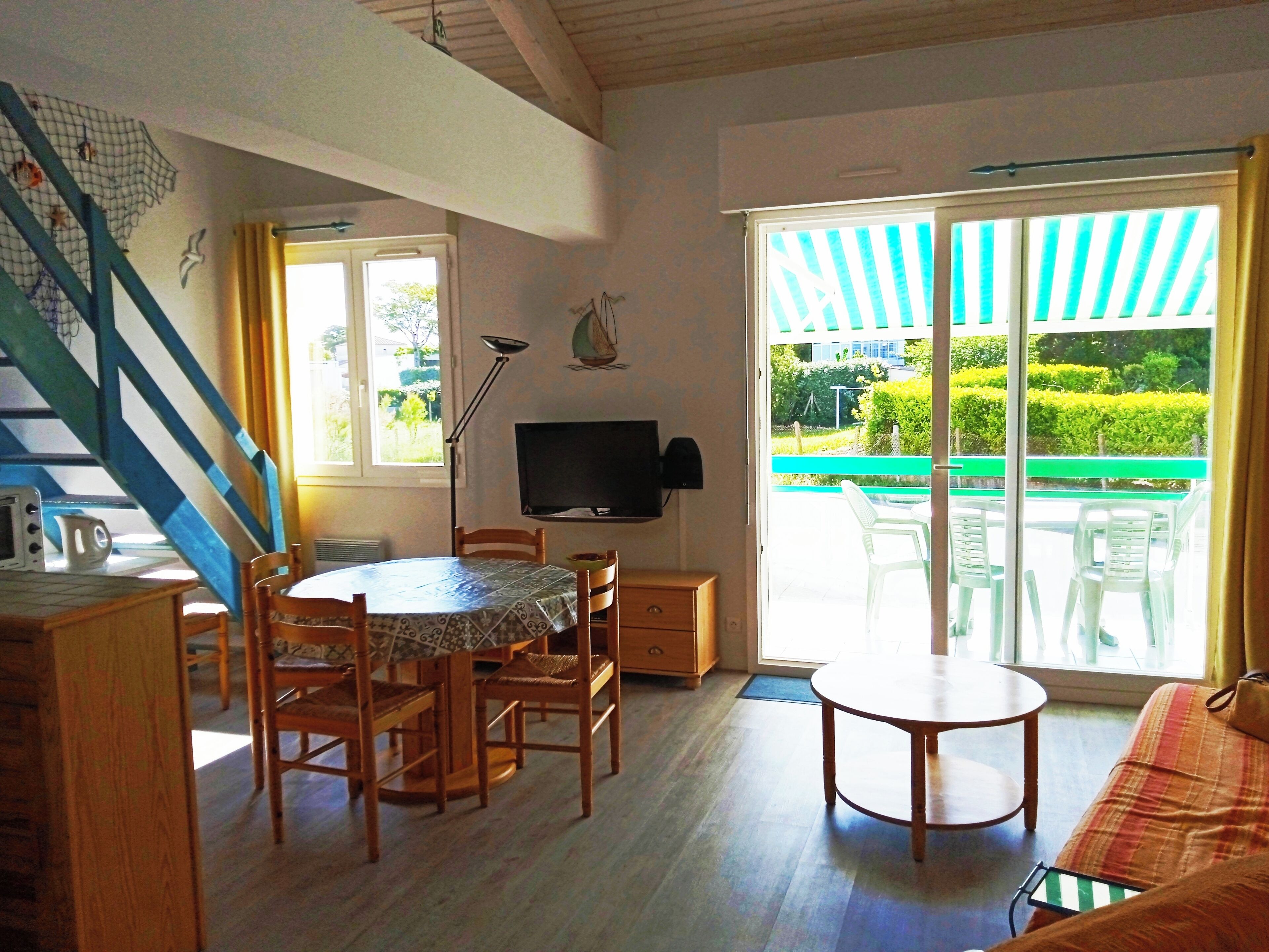 Una habitación con una mesa de comedor, sillas, un televisor y un sofá. Hay una escalera con barandillas azules y vistas a un jardín por la ventana.