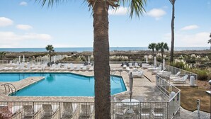 A pool area with lounge chairs and umbrellas, surrounded by palm trees and a view of the beach. - Ocean One 213 - Second Floor Oceanfront 3BR Condo (Hilton Head Island)