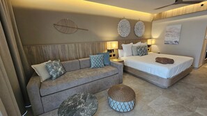 A modern hotel room with a wooden headboard, a sofa with patterned cushions, a bed with a rolled towel, two wall-mounted lamps, and a round woven ottoman. - Serenade Caribe Club Family Beach Resort (Punta Cana)