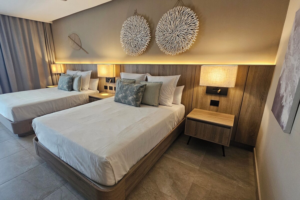 A hotel room with two beds, a wooden headboard, a wall-mounted lamp, and a large window with curtains.