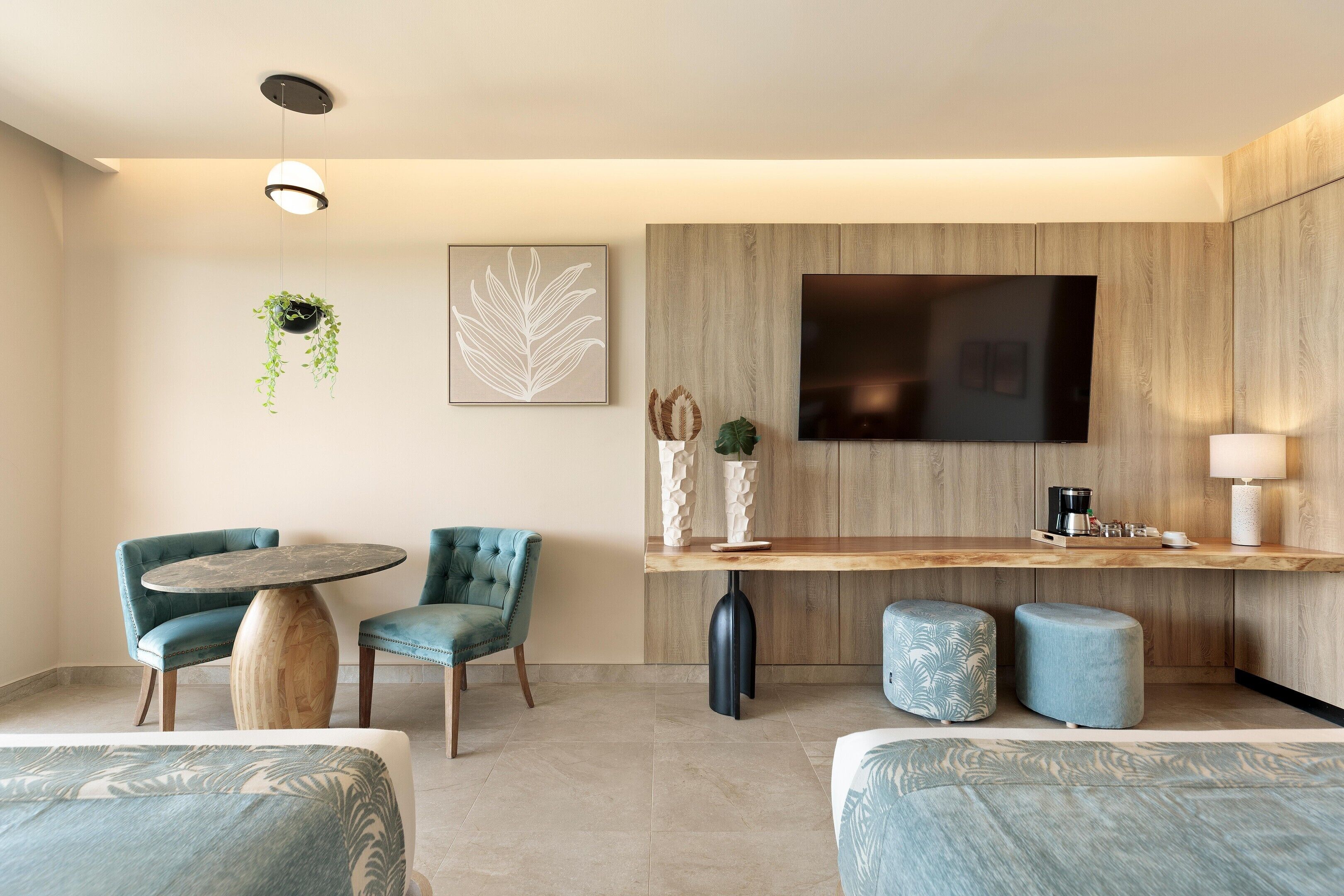 A modern hotel room with a round table, two blue chairs, a sofa, a TV, a coffee machine, and a hanging plant.