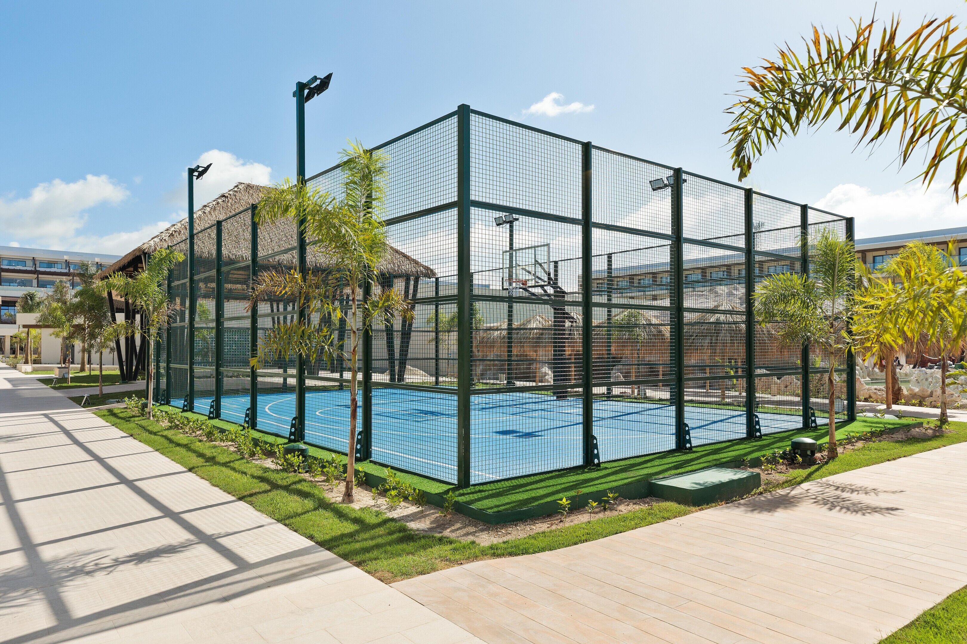 A fenced tennis court with a thatched-roof structure nearby.