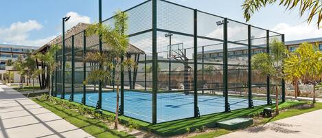 A fenced tennis court with a thatched-roof structure nearby.