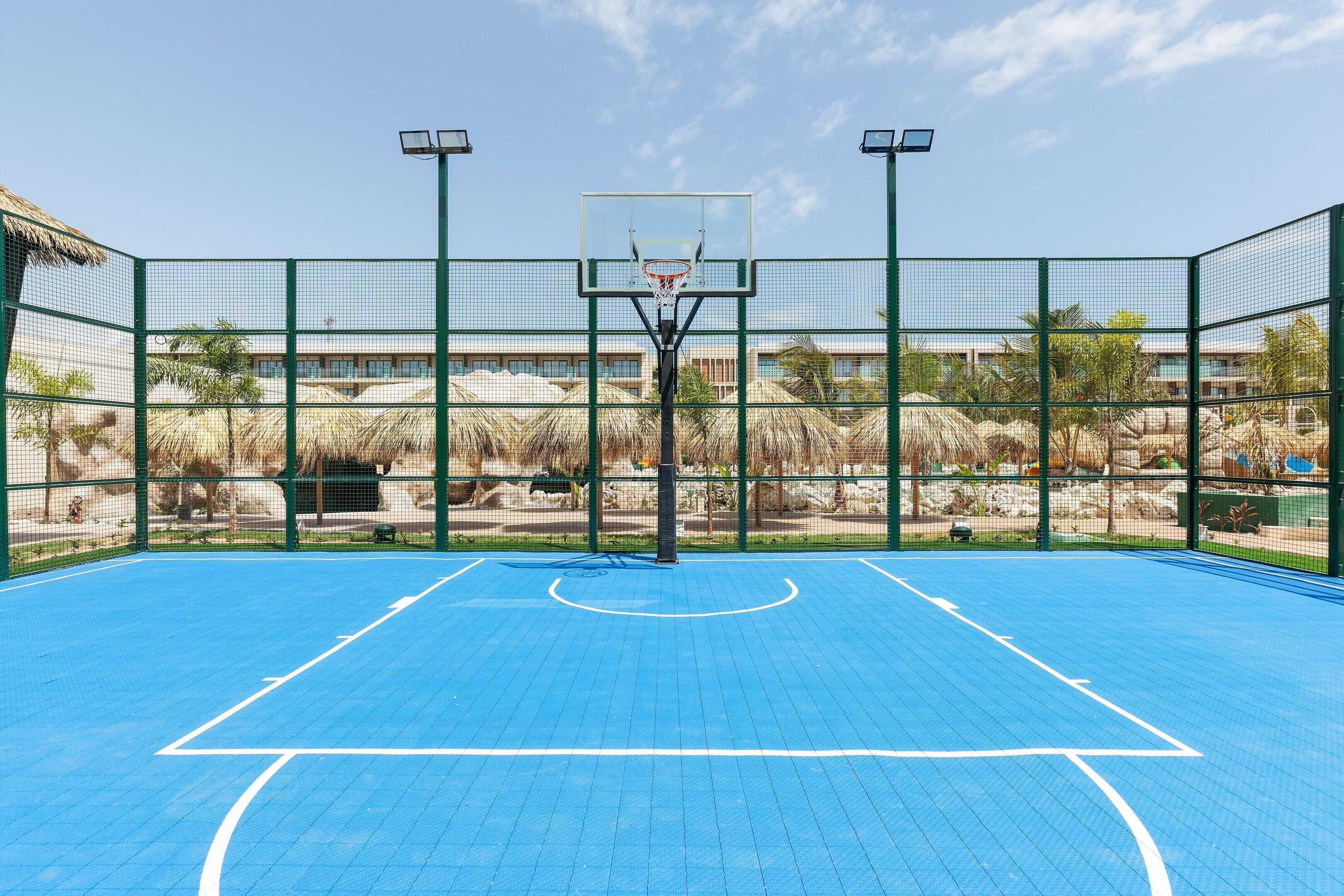 An outdoor basketball court with a green fence, a basketball hoop, and a clear sky.