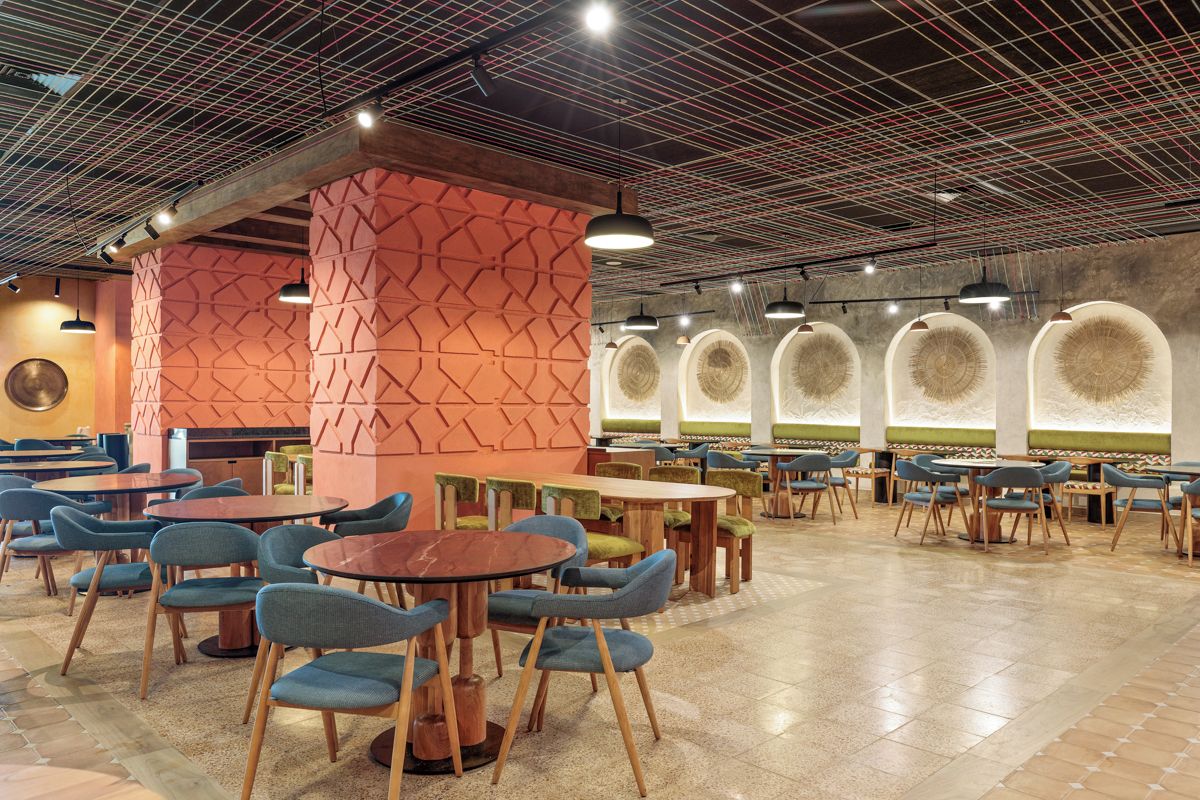 A spacious indoor dining area with multiple tables and chairs, a distinctive geometric wall pattern, and a ceiling with exposed beams and lighting fixtures.