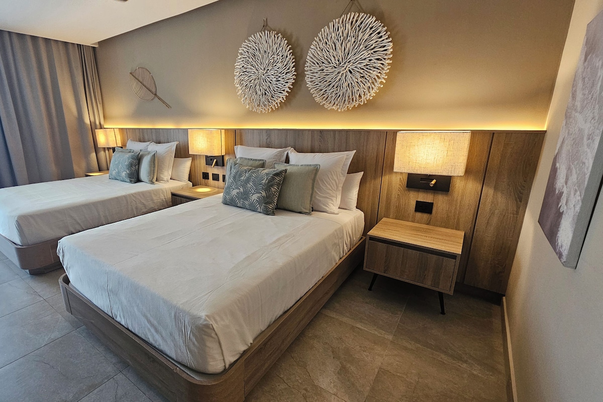 Habitación de hotel con dos camas, cabecera de madera, lámpara de pared y un ventanal con cortinas.