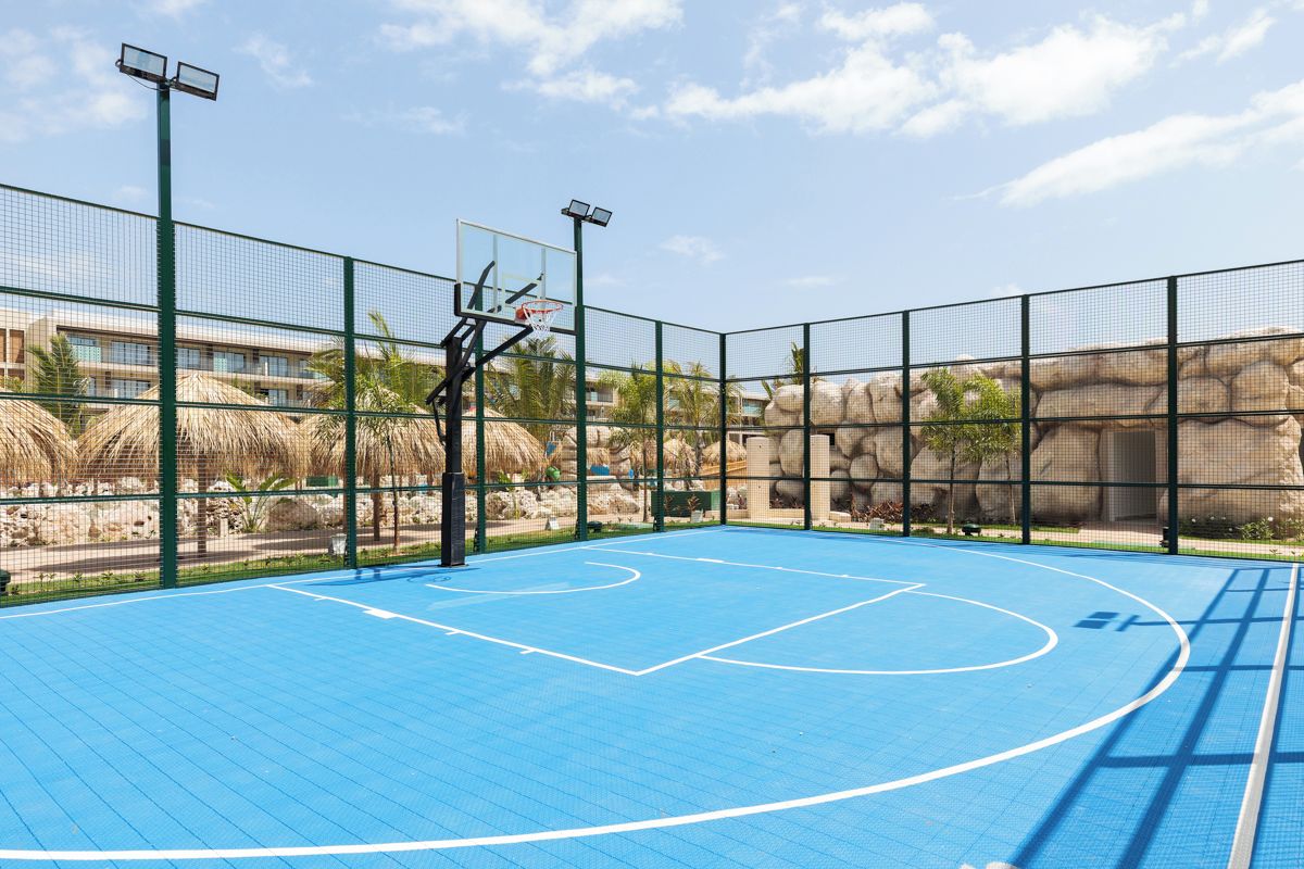 Quadra de basquete externa com cesto, cercada por grade, e um prédio ao fundo.