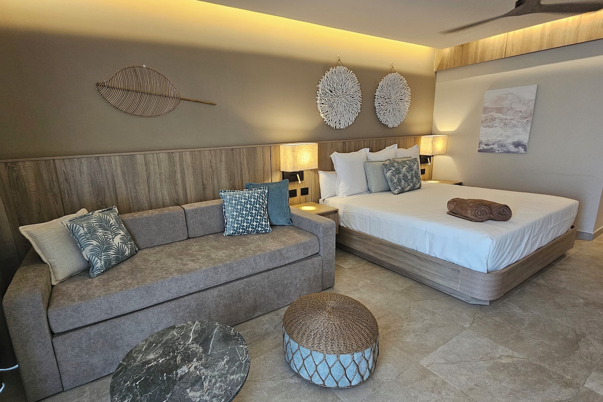 Una habitación de hotel moderna con cabecera de madera, un sofá con cojines estampados, una cama con toalla enrollada, dos lámparas fijadas a la pared y un otoman tejido redondo.