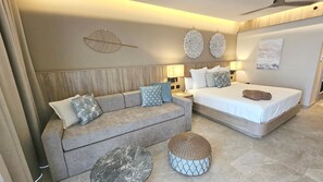 Une chambre d’hôtel moderne comprenant un canapé, un lit, deux tables de chevet, des œuvres d’art murales et un pouf en osier.