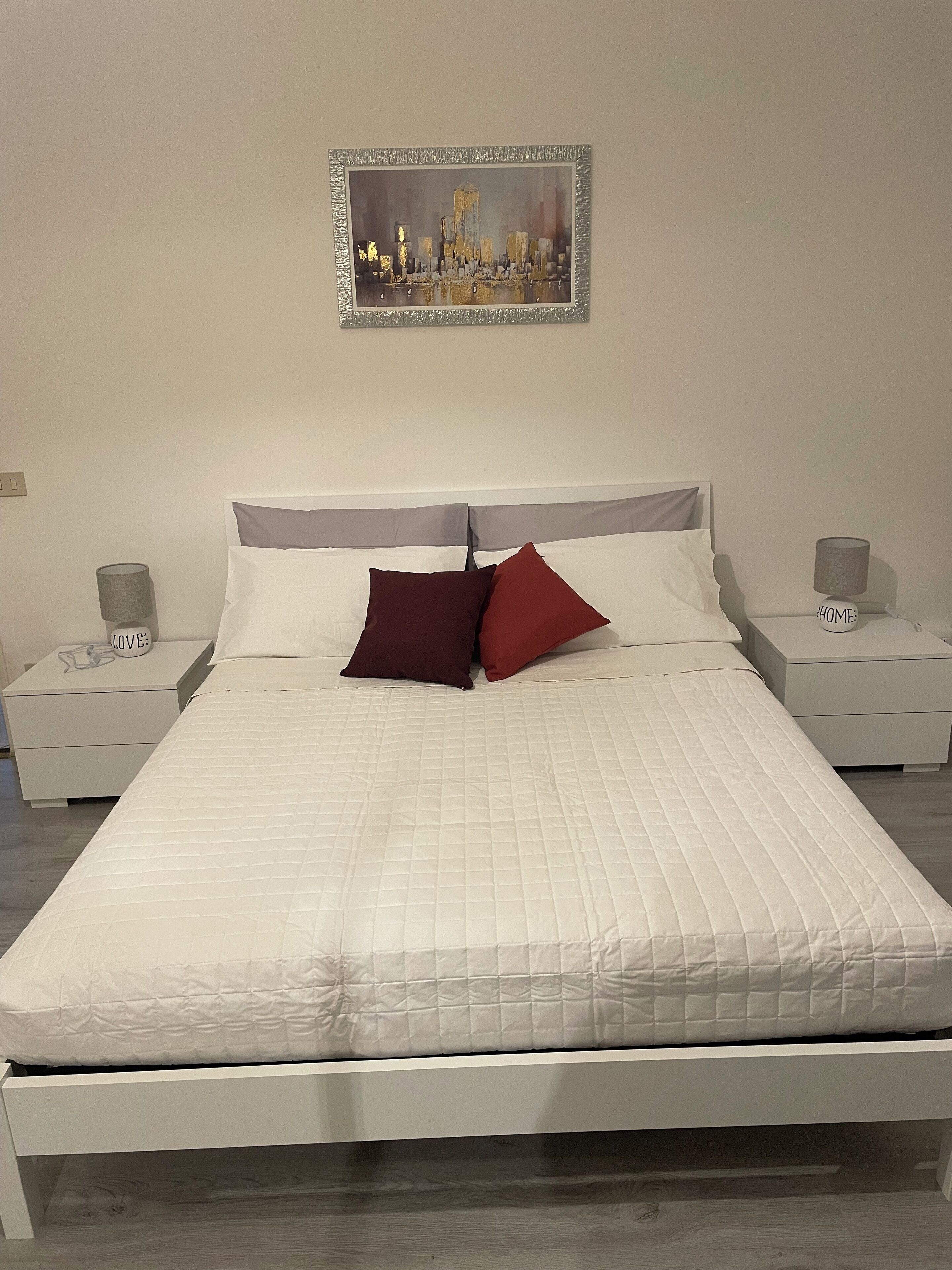 A neatly made bed with white bedding and two red pillows, flanked by white nightstands with lamps and a framed cityscape picture above.