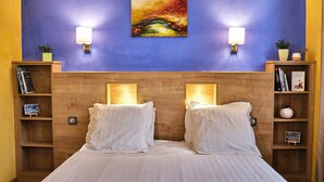 A hotel room with a wooden headboard, two bedside lamps, a painting above the headboard, and bookshelves with books and decorative items.
