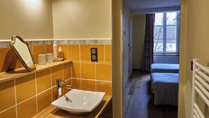 A bathroom with a mirror, sink, and toiletries on a shelf. A bed is visible in the adjacent room.