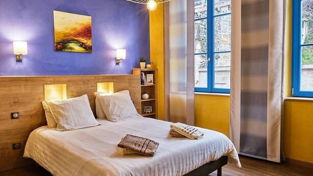 A hotel room with a large bed, a headboard with built-in lighting, a wall-mounted painting, and a window with a view of the outside.