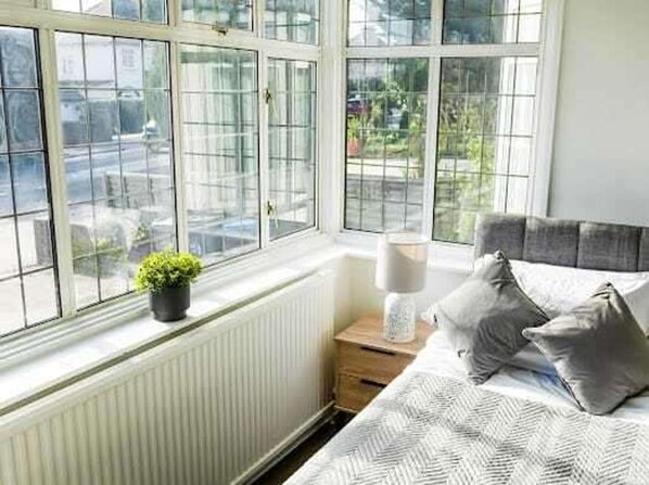 A bedroom with a large window, a bedside table with a lamp, and a bed with pillows. - Serviced Accom Sleeps 8- Near London, Free Parking (South Croydon)