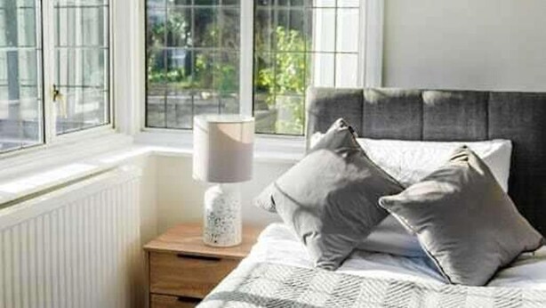 Een slaapkamer met een groot raam, een nachtkastje met een lamp en een bed met kussens.