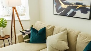 Een beige hoekbank met teal- en witte kussens, een klein bijzettafeltje met een plant en een abstract schilderij in een lijst aan de muur.