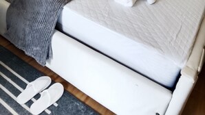 Cama bem arrumada com manta cinza, almofadas brancas e toalha branca enrolada.