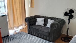 Uma sala de estar com um sofá cinza escuro, um tapete cinza, um ventilador e uma janela com cortinas.