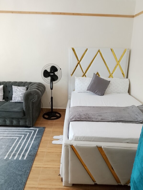 A bedroom with a bed, a grey sofa, a standing fan, and a patterned rug.
